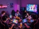 Friends enjoying interactive porn games in a lively gaming lounge filled with neon lights and laptops.